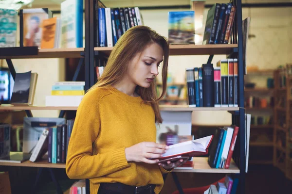 Side view of concentrated female in casual clothes standing near bookshelf reading book while spending free time in light library