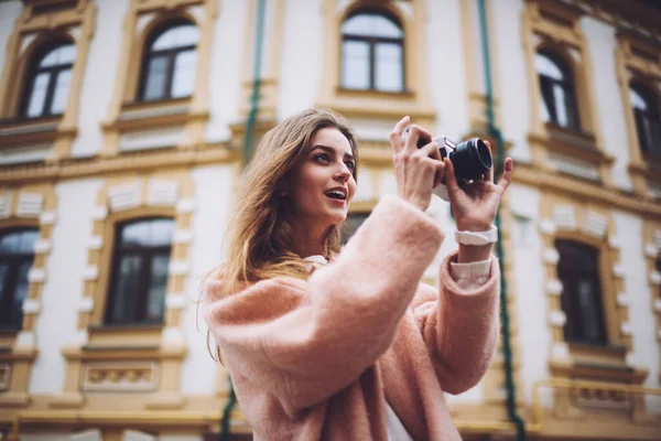 Düşük açılı, pembe hırkalı, gülümseyen kadın fotoğrafçı şehrin eski merkezinde eski bir kamerayla fotoğraf çekiyor.