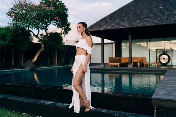 Havuz kenarındaki ahşap terasta tek başına dinlenirken şık giyinmiş ve gözlerini kaçıran çıplak ayaklı turistin yan görüntüsü.