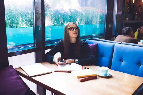 Uzun saçlı, dalgın bir kız öğrenci elinde kahve fincanıyla masaya oturuyor ve cep telefonu kullanırken defteri açıyor.