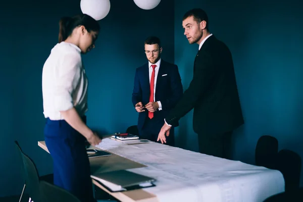 Confident young coworkers standing around table with documents and discussing plan details with each other in modern workplace and wearing elegant formal clothes