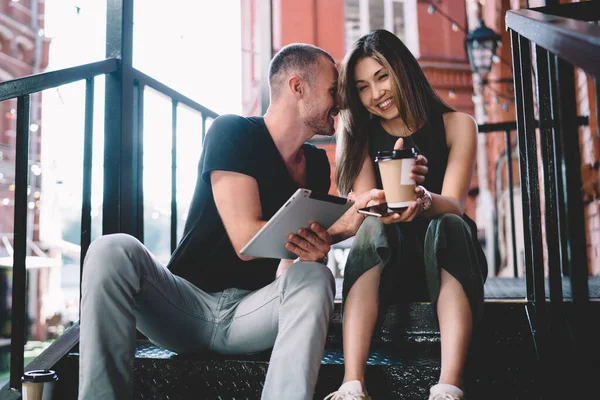 From below of cheerful ethnic couple in casual clothes sitting on stairs and browsing tablet while chilling and drinking takeaway coffee