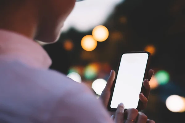 Crop unrecognizable person in eyeglasses checking social media on mobile phone and using internet while standing on city street with bokeh lights on background