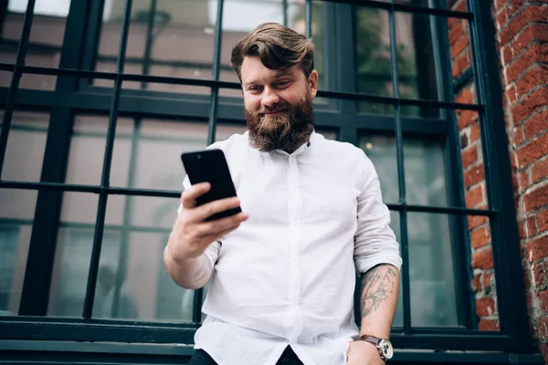 Low angle of positive bearded male entrepreneur with hand in pocket browsing mobile phone while standing near brick building and looking down