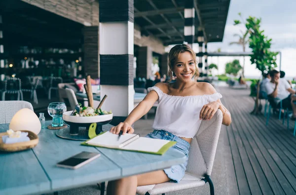 Smiling young female in trendy outfit sitting at table with opened notebook and looking at camera happily in modern spacious restaurant on terrace