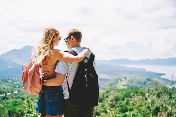 Back view of anonymous young couple of tourists enjoying nature while standing on hill and hugging each other while wearing summer clothes