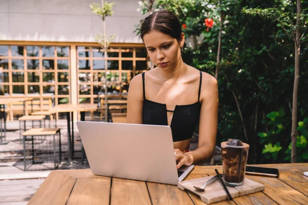 Young female freelancer in trendy outfit sitting at table with cold drink and smartfon while typing on laptop during remote work
