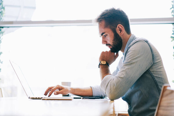 Side view of bearded man in casual outfit holding hand at chin sitting at table near window and working on laptop