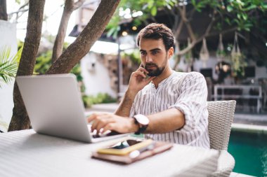Yoğunlaşmış genç yakışıklı sakallı iş adamı, yazın havuz başında bulanık bir arka plan üzerinde netbook 'ta verileri kontrol ediyor.
