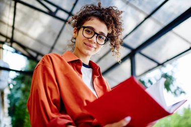 Gözlükleriyle kameraya bakan iyimser etnik kız öğrenci ders kitabında bilgi okurken sokaktaki üniversite için proje yapıyor.
