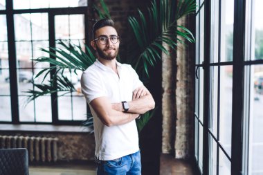 Klasik optik gözlüklü Türk hippinin yarı boy portresi kameraya bakarak ve içeride poz vererek görüş koruması sağlıyor.