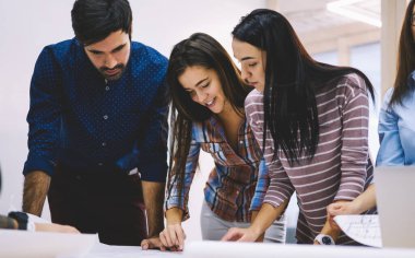 Bir grup gülümseyen iş arkadaşı masanın etrafında toplanır ve iş yerindeki projenin detaylarını tartışırken birbirlerine yakın dururlar.