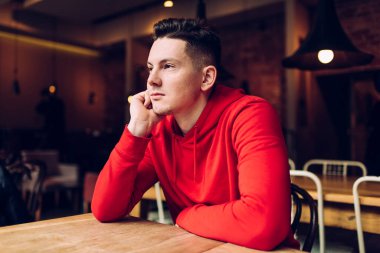 Kırmızı kapüşonlu, ciddi, genç siyah erkek. Kafede el pençe divan oturuyor ve dalgın dalgın bakıyor.