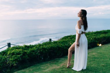 Uzun boylu, çıplak ayaklı, beyaz elbiseli, yeşil çimlerin üzerinde duran ve muhteşem manzaranın tadını çıkaran, şehvetli bir kadın.