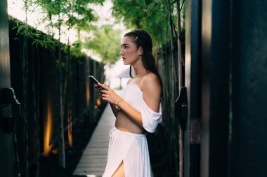 Metal kapıların yanında duran odaklanmış bir kadının yan görüntüsü ve tropik tatil beldesinde dinlenirken cep telefonunda mesajlaşması