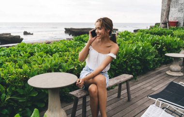 Yeşil bitkilerin yanındaki bankta oturan ve tatil köyünde dinlenirken cep telefonuyla konuşan mutlu, genç bir kadın.