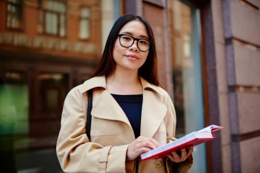 Sokakta durup not defterindeki notları okurken, gözlüklü, sıradan giyinen, efkârlı bir etnik kız öğrenci.