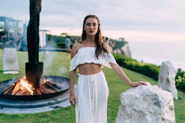 Şenlik ateşinin yanında duran, taşa yaslanan ve tatil köyünde vakit geçirirken kameraya bakan tatmin olmuş genç kadın.