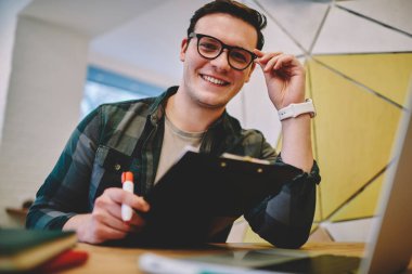 Düşük açılı, dişlek tebessümlü, kameraya bakan, gözlüklü ve pano kullanarak masada oturan pozitif bir adam. 