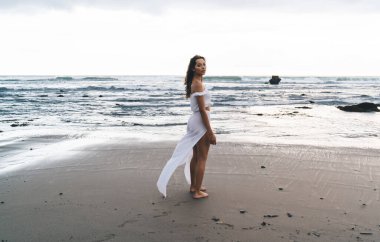 Yazlık elbiseli, kumlu deniz kıyısında bulutlu gökyüzüne bakarken kameraya bakan sakin, genç, etnik bir kadın gezgin.