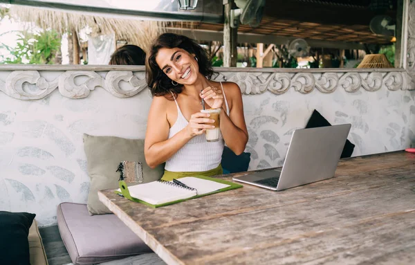 Young content ethnic distance employee with glass of latte macchiato sitting at cafeteria table with netbook and journal while looking at camera on terrace