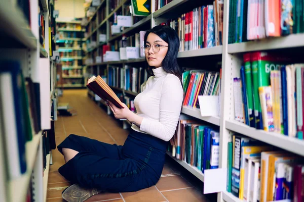 亚洲黑发女子 戴着白色高领眼镜 蓝色长裤 双腿交叉地坐在书架旁边 翻开书本 — 图库照片