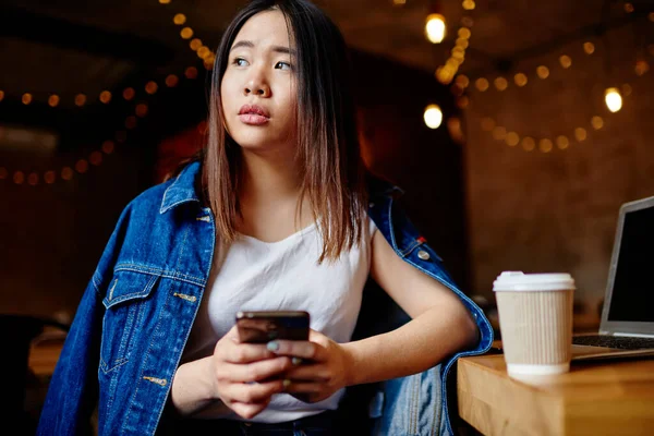 Serious Asian woman in casual clothes with takeaway coffee looking away and using mobile phone for chatting during break in cafe