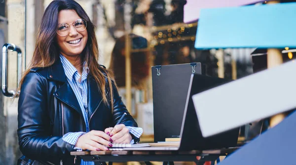 Female with toothy smile in casual outfit looking away sitting at table while using laptop for work in cafe on street