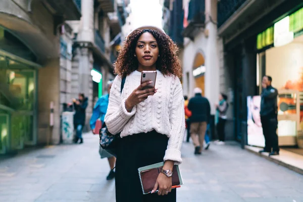 African American woman in casual wear using phone to type text message while walking around city and looking at camera