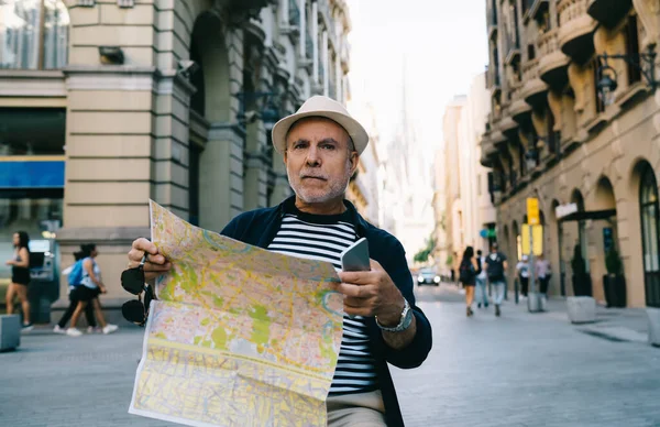 Adult man in straw hat holding cellphone device and location paper in hands thinking about direction for exploring travel town on holidays