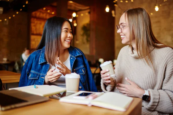 Glad Asian woman with toothy smile and cheerful female friend with takeaway coffee sitting at table in cafe having conversation