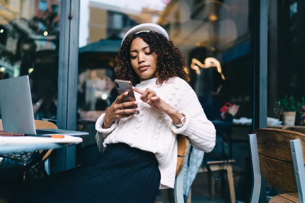 Skilled dark skinned woman freelancer working on cafe terrace holding mobile phone connected to 4G