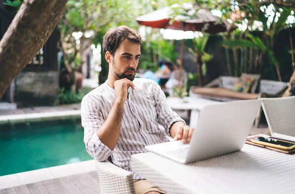 Ciddi, sakallı genç bir erkek havuz kenarındaki masada bilgi topluyor. Gündüz vakti arka planda bulanıklık var.