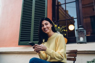 Sokak terasında tahta koltukta dinlenen düz siyah saçlı ve cep telefonuyla mesajlaşan genç etnik kadın.