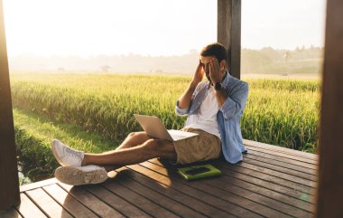 Yorgun, uzaktan kumandalı bir erkek işçi, renkli tarlanın yanındaki ahşap terasta bacak bacak bacak üstüne atarak otururken kafasına dokunuyor.