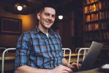 Samimi kütüphanede lamba ışığında çalışırken ve kameraya gülümserken kulaklık takan genç ve mutlu bir erkek.