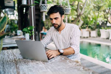 Havuza karşı tropikal tatil beldesi terasında otururken şık gömlekli sakallı genç erkek netbook okuyor.