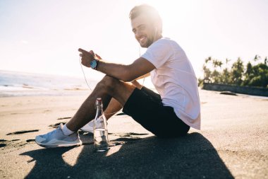 Sahil kıyısında bir şişe suyla otururken kulaklıkla müzik dinleyen spor giyimli mutlu bir erkeğin alçak açılı görüntüsü.
