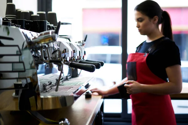 Confident female waiter in uniform using professional italian coffee machine for preparing order in cafeteria