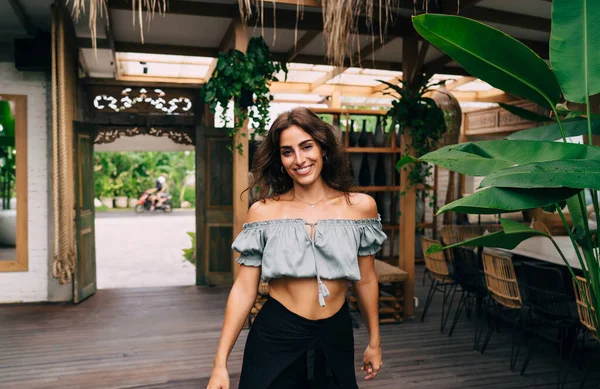 Satisfied ethnic female in stylish outfit standing in cafe near green plant in summer day and looking at camera with toothy smile