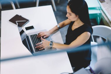 Yukarıdan, planlamacı ve akıllı telefonla masada oturan ve bilgi aramak için dizüstü bilgisayarını kullanan genç bir kadın işçi geliyor.