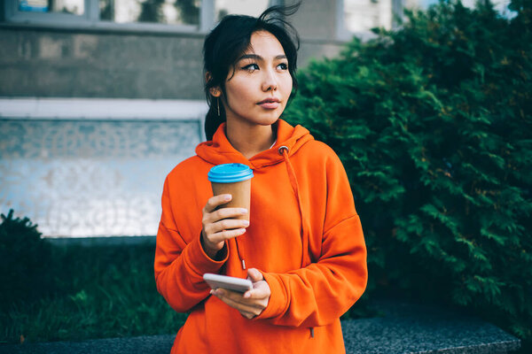 Thoughtful Asian female in vivid outfit looking away while standing on street with coffee and using mobile phone for browsing internet