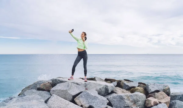 携帯電話で手と挨拶の友人を振っている間 スポーツウェア姿の若い女性が石の上に立ち 海岸で自画自慰行為 — ストック写真