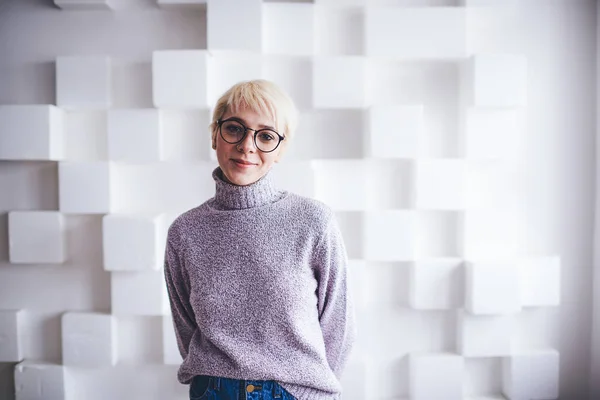 Young positive female in casual clothes with eyeglasses and blond hair looking at camera standing on white background with hands behind back