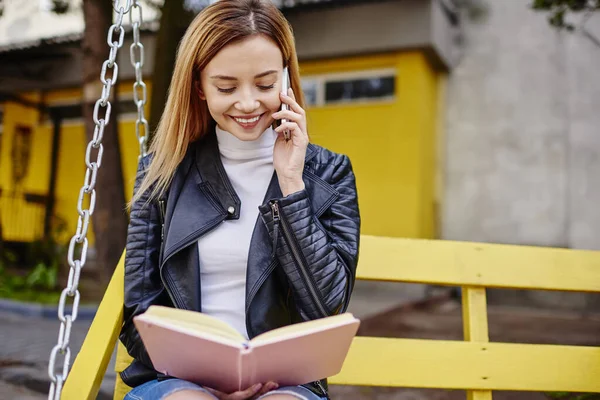 Siyah deri ceketli neşeli bir bayan parkta salıncakta oturuyor ve telefonda konuşurken günlükte notlar okuyor.