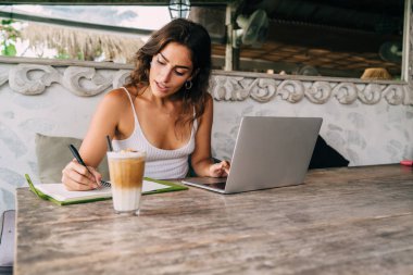 Genç odaklı etnik bayan uzaktan çalışan netbook ve cafe terasındaki bir bardak latte macchiato 'nun yanındaki günlüğe not alıyor.