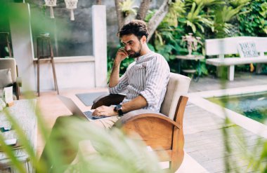 Rahat koltukta otururken internette sörf yapmak için dizüstü bilgisayar kullanan rahat sakallı bir adamın yan görüntüsü.