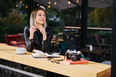 Kafeteryada elinde bir bardak içki ve fotoğraf makinesiyle masada oturan gülümseyen genç bayan. 