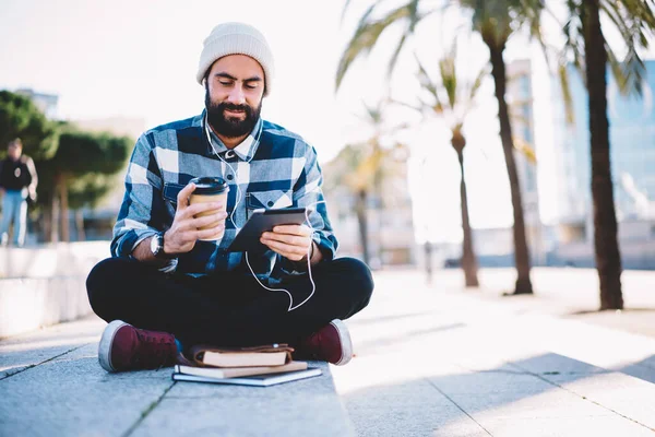 Middle Eastern hipster guy in electronic headphones using touch pad technology holding takeaway cup