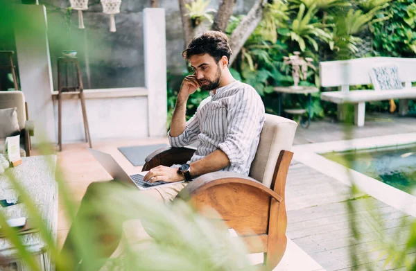 Rahat koltukta otururken internette sörf yapmak için dizüstü bilgisayar kullanan rahat sakallı bir adamın yan görüntüsü.